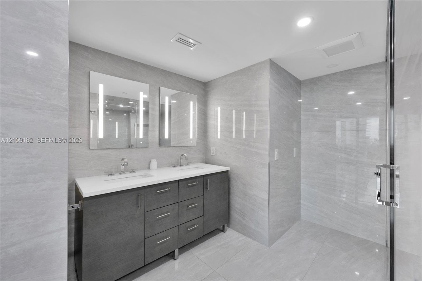 Bathroom featuring a dual-sink vanity with a white countertop, large-format grey stone-patterned wall tile, polished chrome fixtures, and dual integrated LED-backlit mirrors