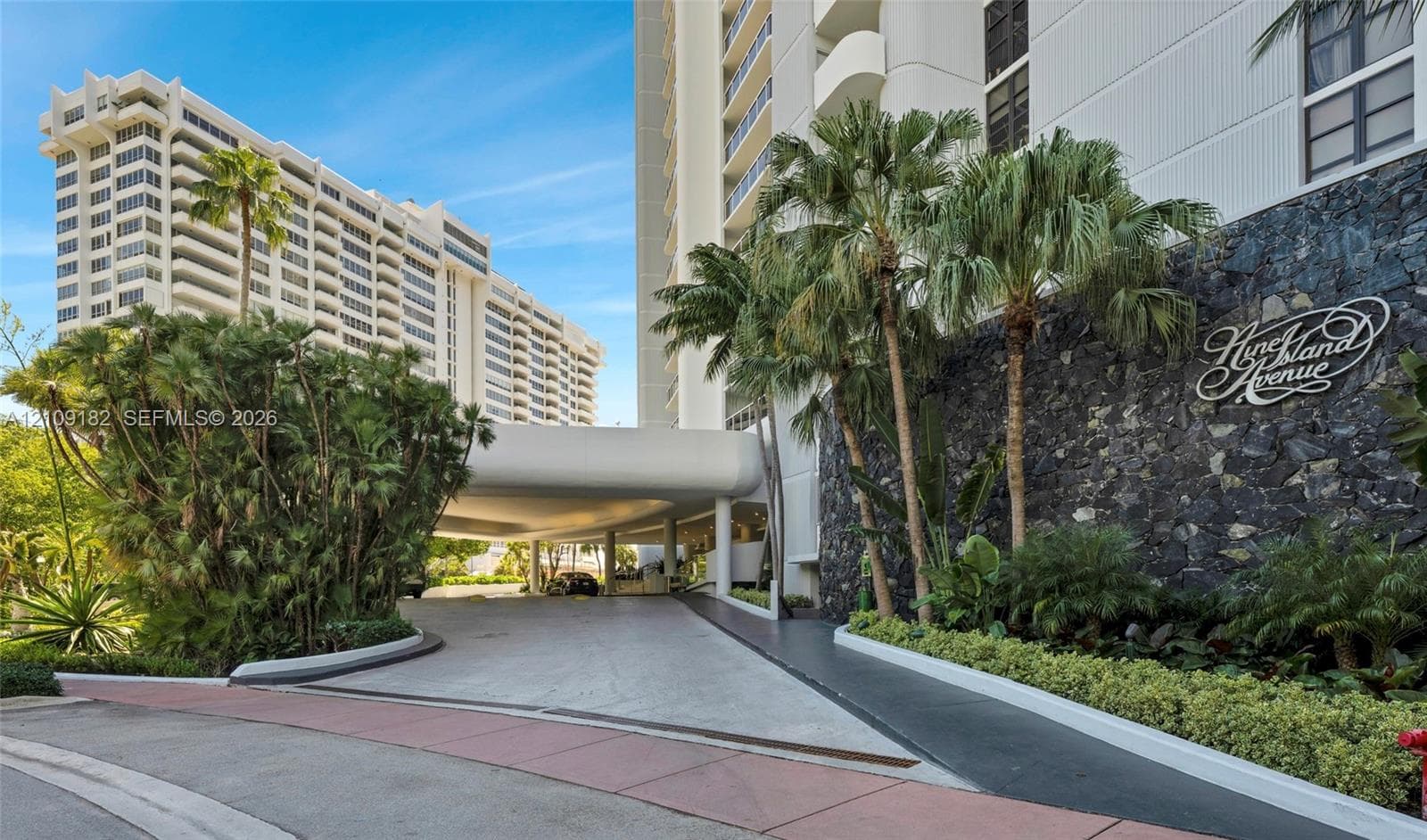 Multi-story high-rise exterior featuring a concrete porte-cochère, decorative stone masonry wall, mature palm trees, and landscaped perimeter hedging