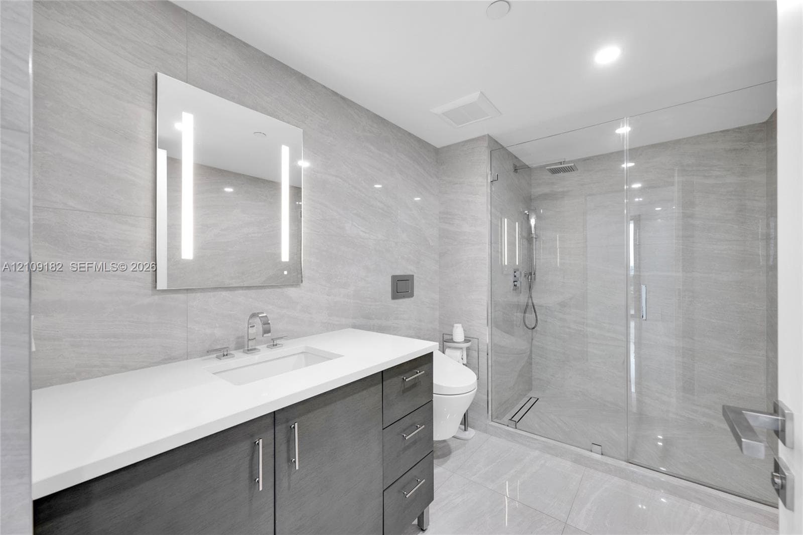 Modern vanity with stone countertop and integrated sink, full-height stone wall tiling, LED-backlit mirror, glass-enclosed shower with rainfall showerhead, and wall-mounted toilet
