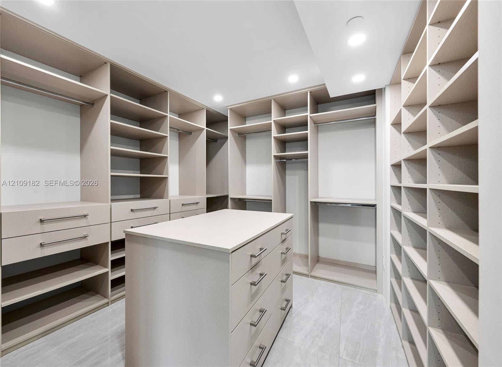 Custom walk-in closet featuring extensive built-in shelving, multiple hanging rods, integrated storage drawers, and a central island with a stone-finish countertop over tiled flooring