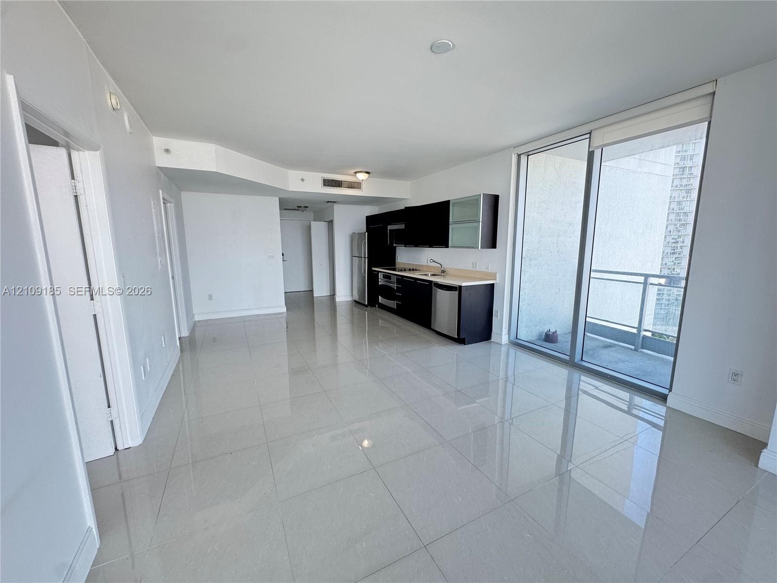 Open-concept interior featuring large-format tile flooring, cabinetry with dark finishes, stone-surface countertops, stainless steel appliances, and floor-to-ceiling glass sliding doors opening to a balcony