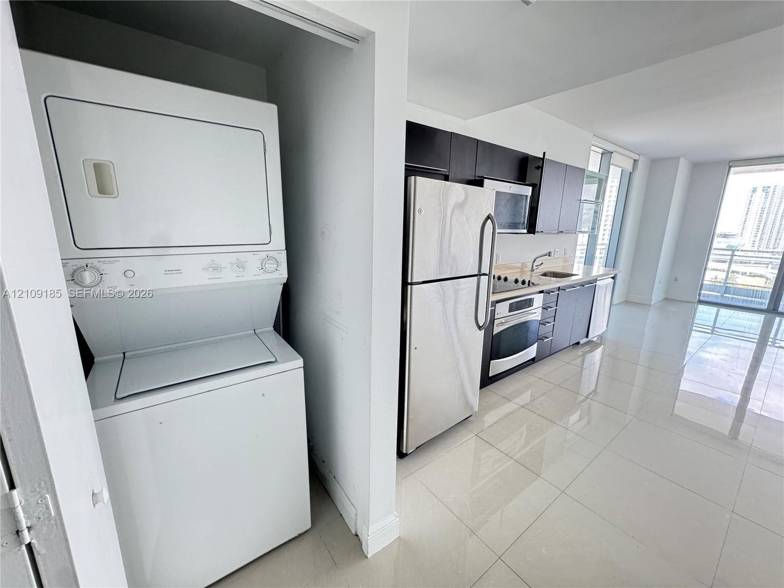 Stacked laundry unit set within a dedicated closet, featuring a kitchen equipped with stainless steel appliances, dark cabinetry, and polished tile flooring throughout