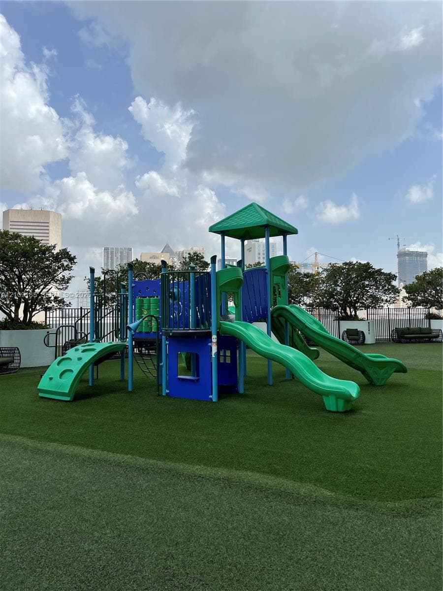 Recreational area featuring a multi-component play structure with dual slides and climbing elements, set upon a synthetic grass surface and enclosed by perimeter fencing