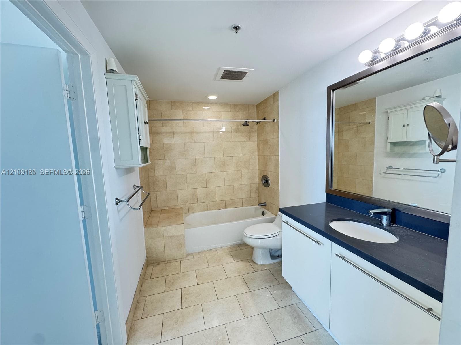 Bathroom featuring white cabinetry, dark countertop with an integrated oval sink, full-width mirror with integrated lighting, and a tile-surround shower and tub combination