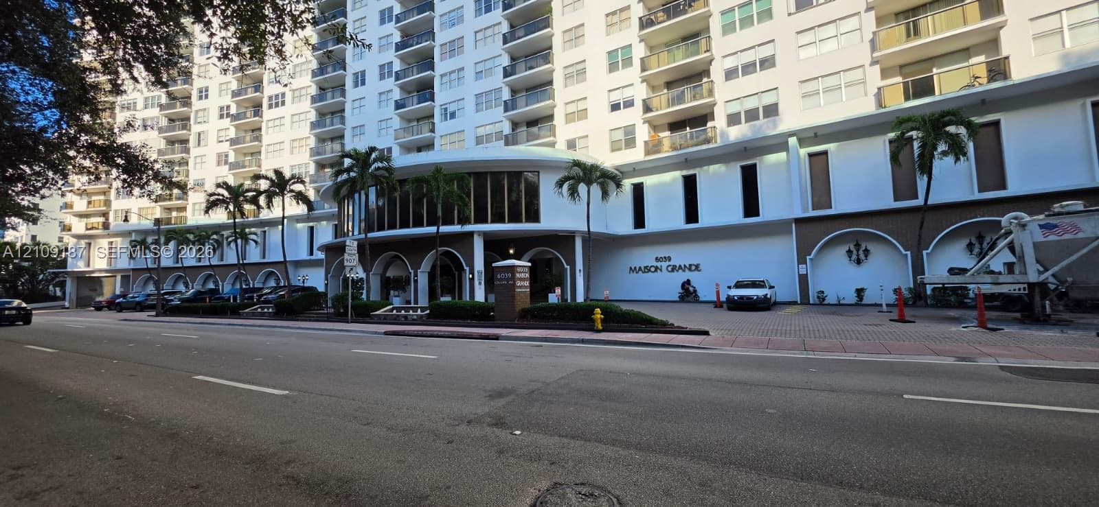 Mid-rise residential building exterior featuring a porte-cochère entry, arched decorative wall niches, multi-story balconies, and lush palm tree landscaping