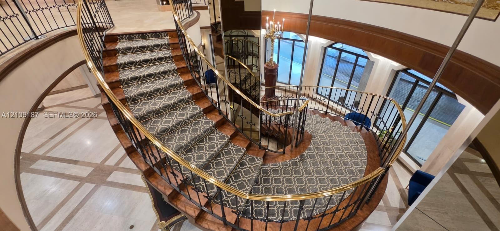 Curved staircase featuring wood-finish treads, custom patterned carpet inlay, wrought iron balusters, and a brass handrail, complemented by stone tile flooring with decorative inlay
