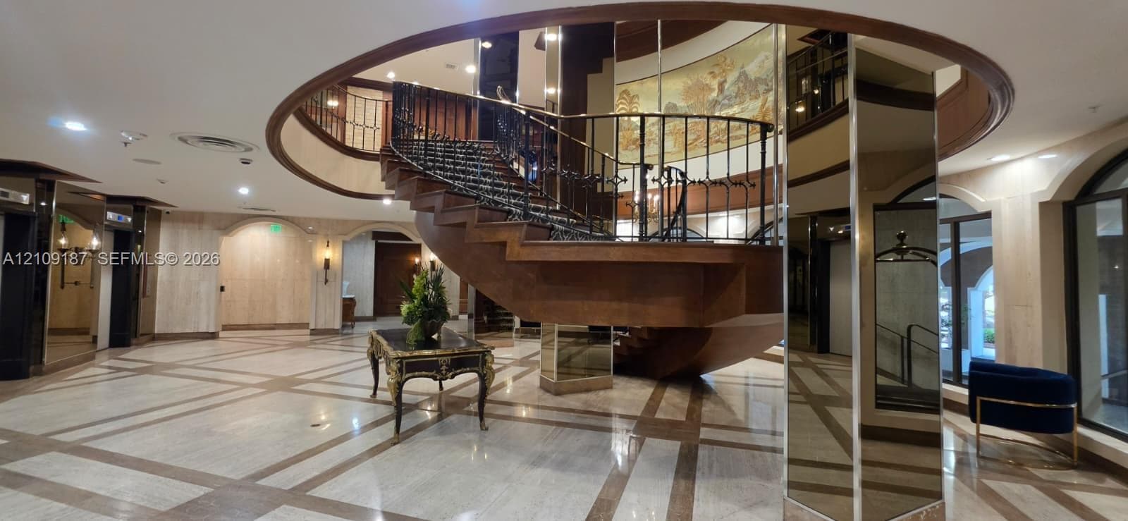 Grand foyer featuring a circular architectural cutout, a sweeping wooden staircase with a metal balustrade, patterned stone flooring, and mirrored support columns