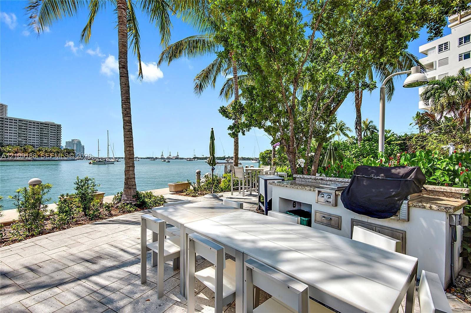 Waterfront patio featuring stone-tiled flooring, a built-in outdoor kitchen with stone countertops and a stainless-steel grill, and mature palm trees