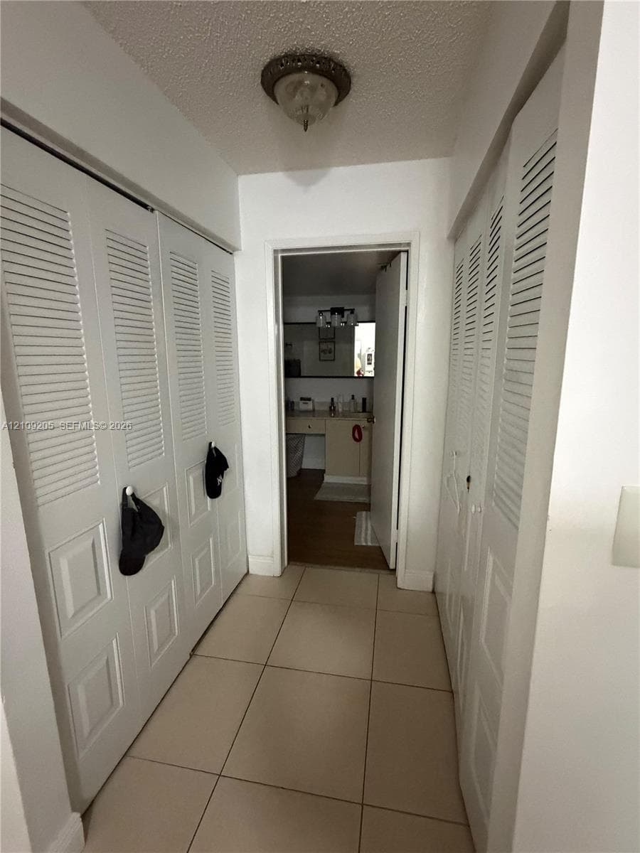 Hallway featuring large-format tile flooring and dual sets of white louvered-panel bi-fold closet doors, illuminated by a ceiling-mounted flush-mount light fixture