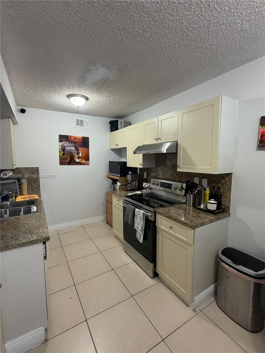 Kitchen featuring cream-toned shaker cabinetry, stone-finish countertops, stainless steel range with overhead hood, and tile flooring