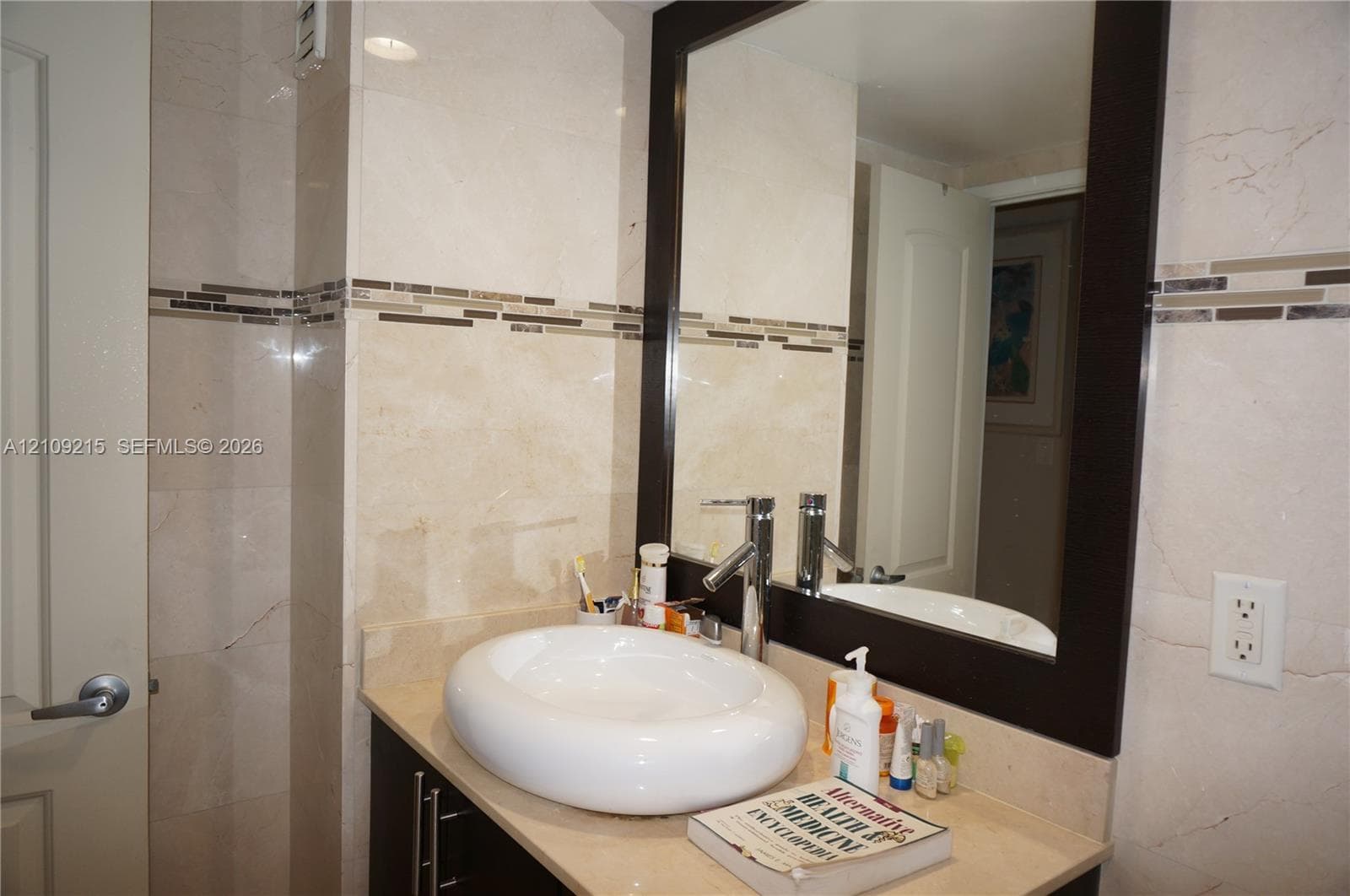 Bathroom vanity featuring a rounded vessel sink, stone-surface countertop, custom dark-finish cabinetry, a framed mirror, and horizontal decorative glass tile backsplash