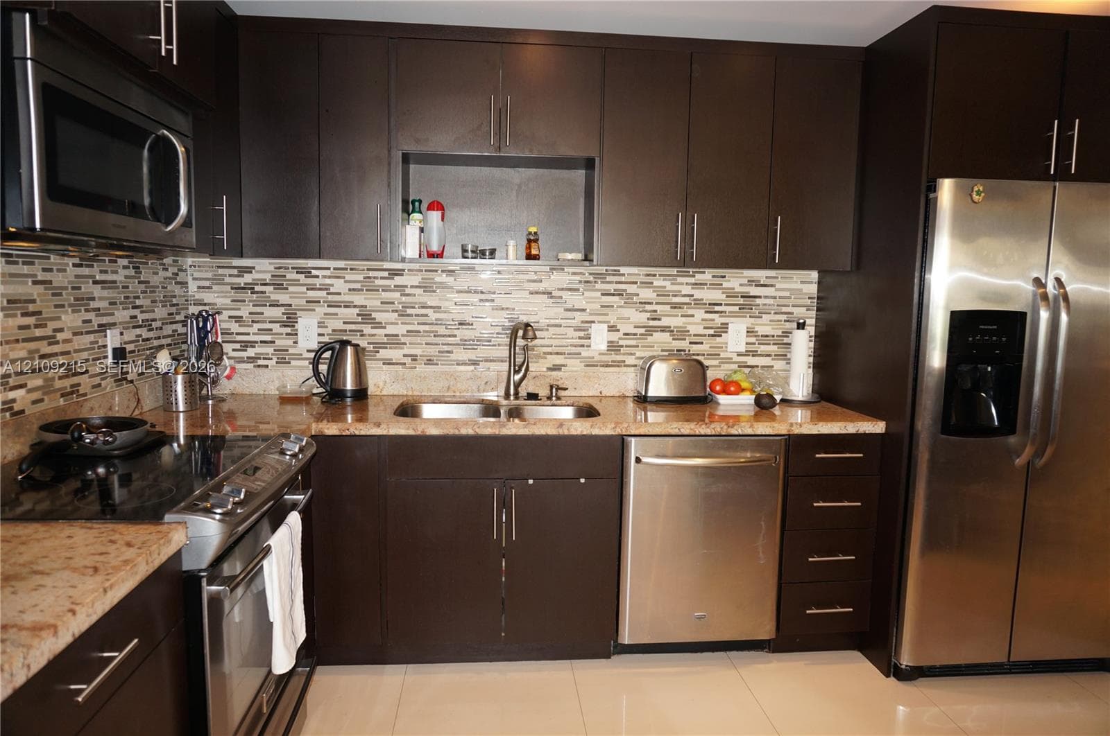 Dark espresso cabinetry with brushed metal pulls, stone countertops, horizontal multi-tonal backsplash, stainless steel appliances, and large-format tile flooring