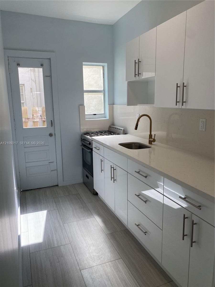 Kitchen featuring white flat-panel cabinetry, light-toned stone-finish countertops, brass-finish faucet, and wood-finish flooring