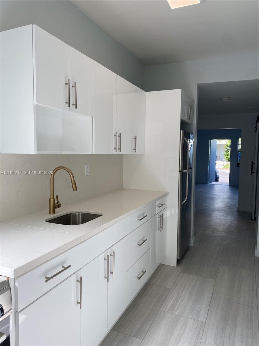 Kitchen featuring white high-gloss cabinetry, solid surface countertops, a stainless steel undermount sink with a gold-tone faucet, and large-format tile flooring