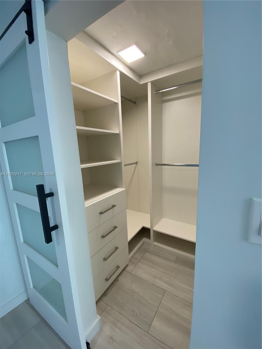 Walk-in closet featuring white built-in shelving, integrated dresser drawers with bar pulls, polished metal hanging rods, frosted glass barn door, and wood-finish flooring