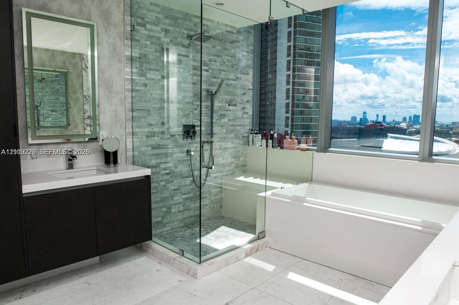 Modern floating vanity with a stone countertop, floor-to-ceiling glass-enclosed shower featuring stone wall tile and an integrated bench, deep soaking tub, and expansive windows showcasing urban scenery