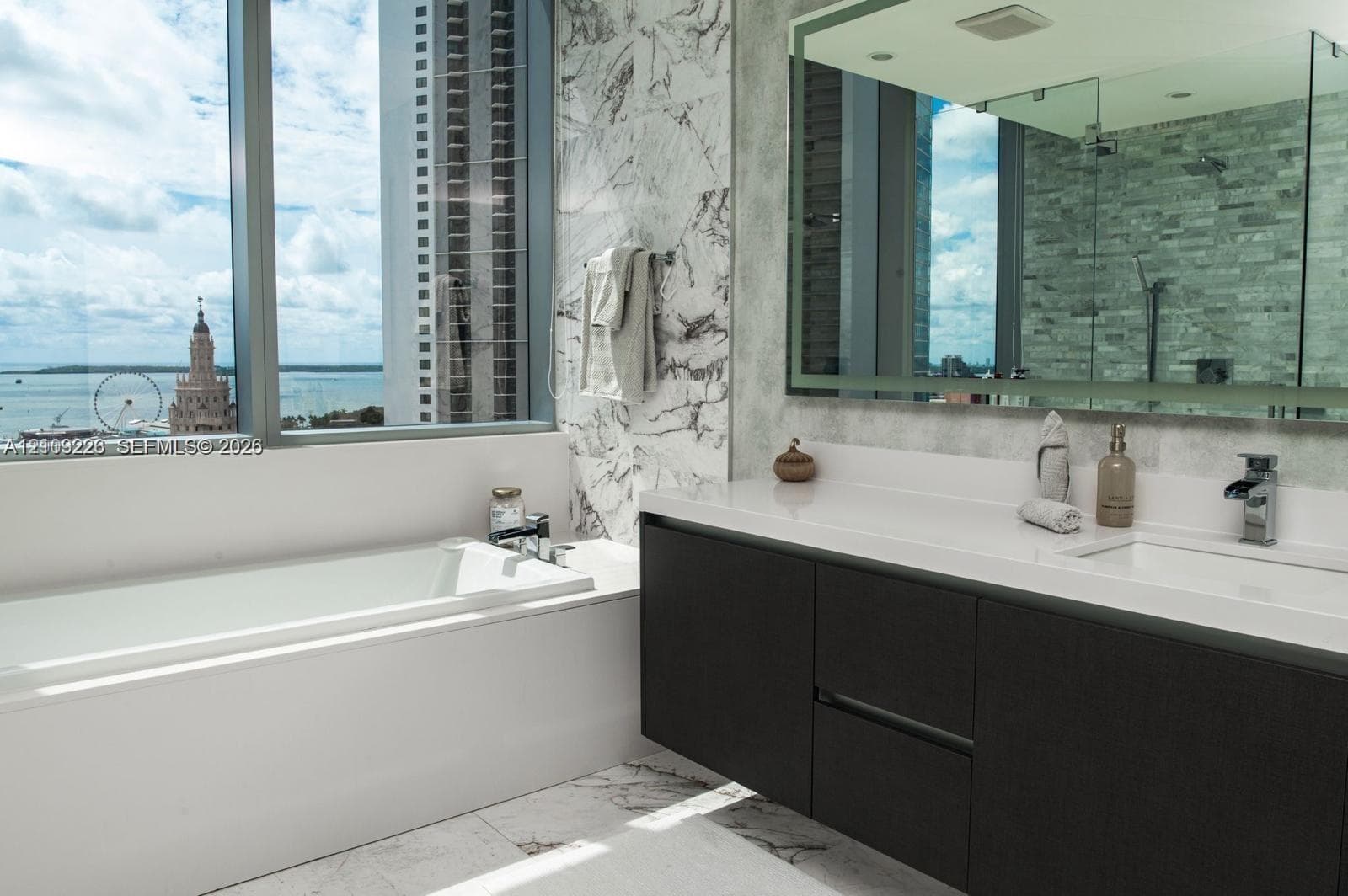 Floating dark cabinetry with an integrated countertop and sink, wall-spanning vanity mirror, floor-to-ceiling stone wall cladding, deep soaking tub, and large-format window with water backdrop