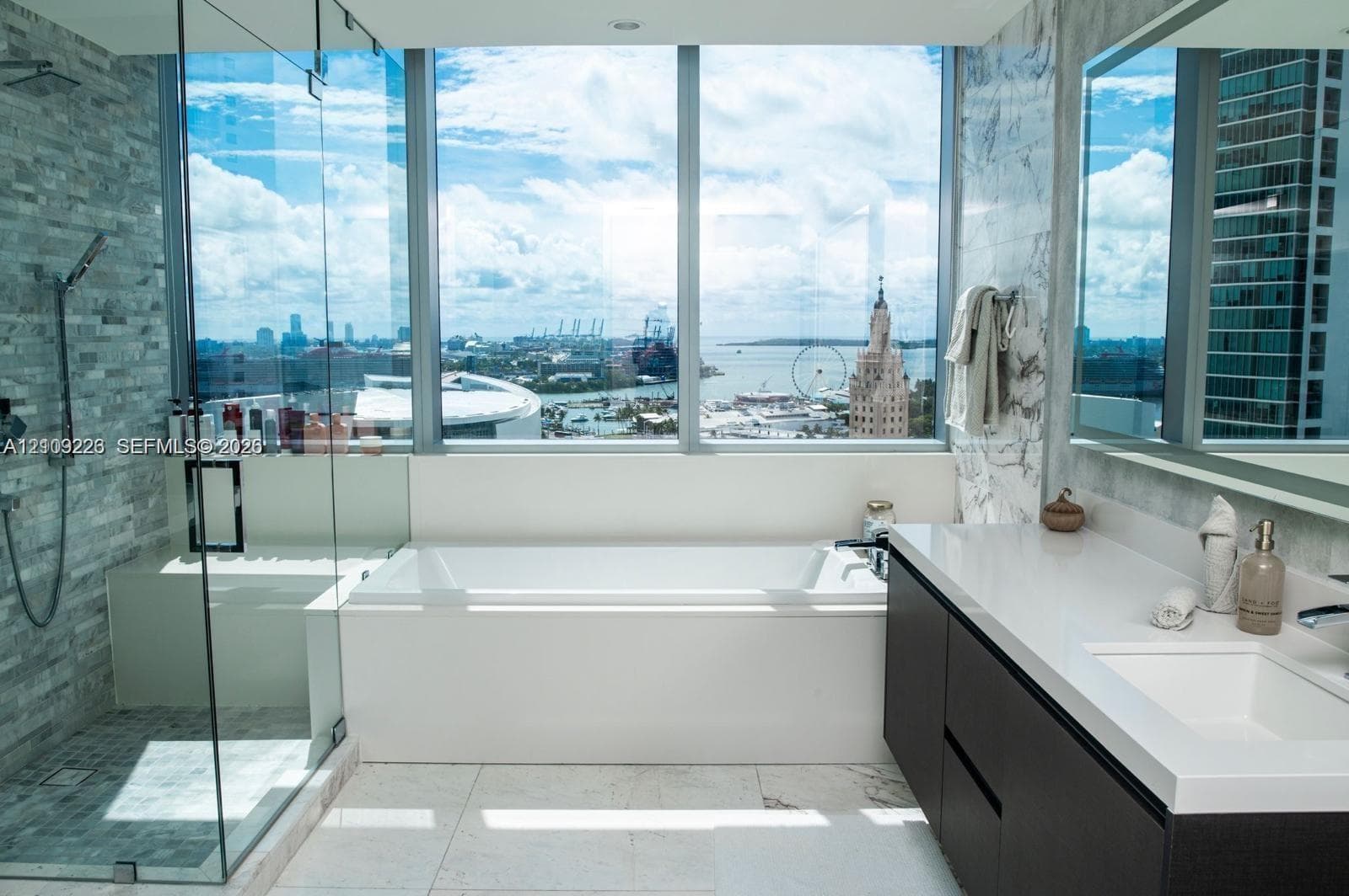 Floor-to-ceiling windows, glass-enclosed walk-in shower with stone tile wall, built-in soaking tub, and dark wood-finish vanity with white countertop