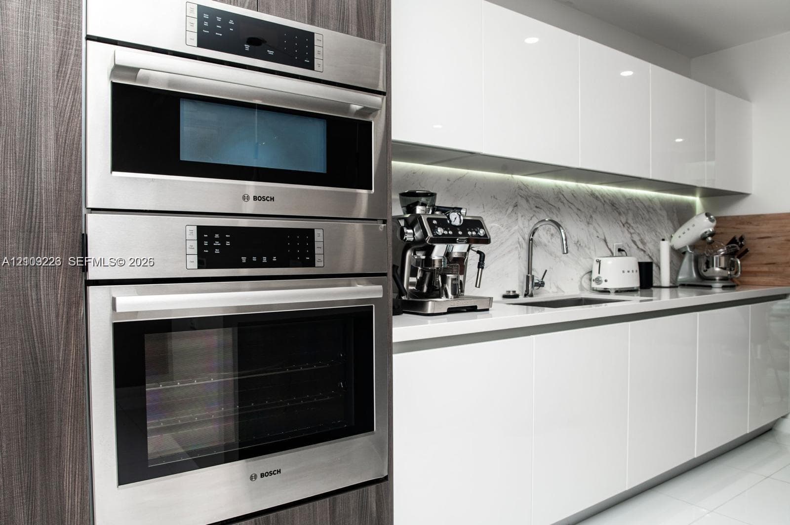 Modern kitchen featuring a stacked stainless steel Bosch wall oven and microwave, high-gloss white cabinetry, stone-finish backsplash, and an undermount sink with a gooseneck faucet