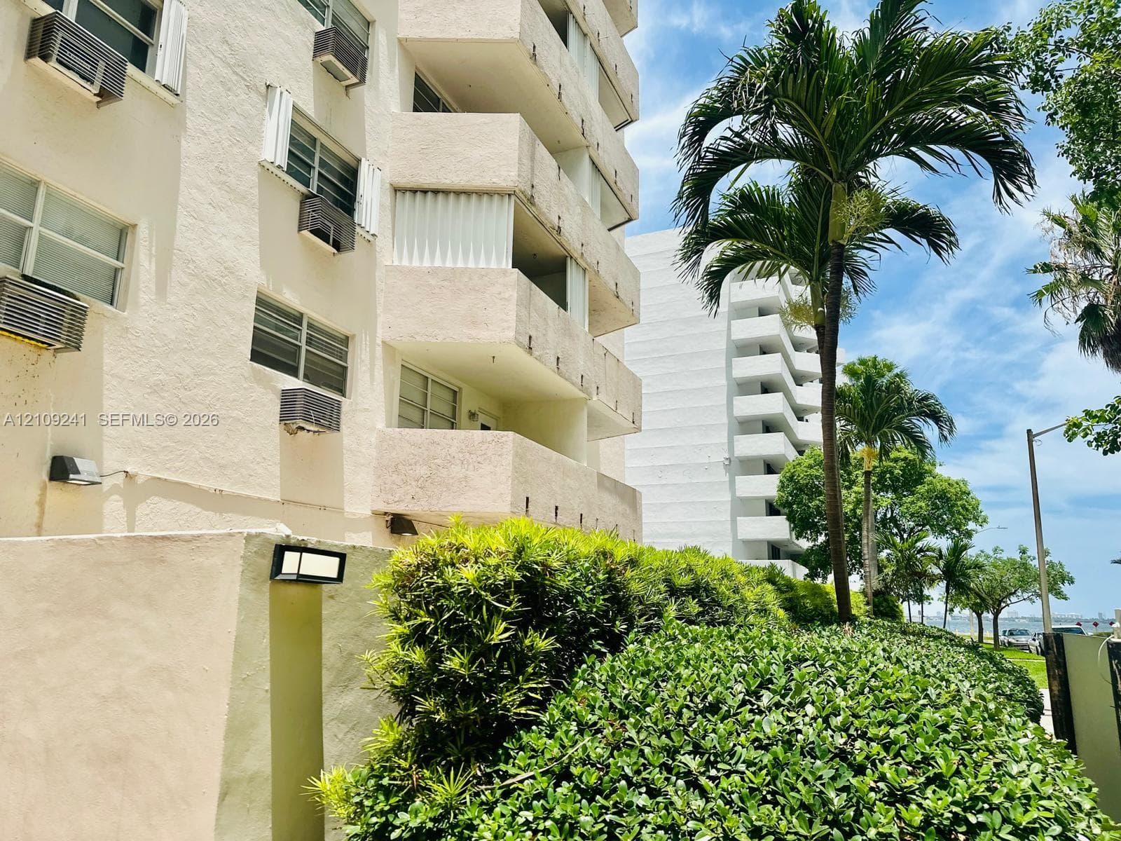 Multi-story residential building exterior featuring stacked private balconies, stucco facade, manicured shrubbery, and mature palm trees