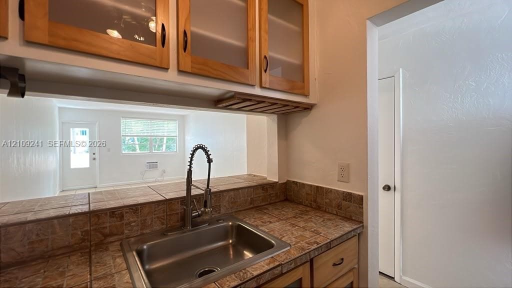 Kitchen area featuring wood cabinetry with glass-panel inserts, built-in stemware rack, tile countertops, and a stainless steel undermount sink with a gooseneck faucet