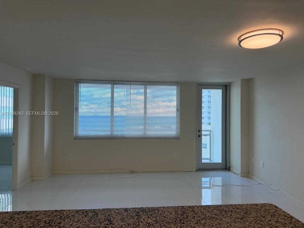 Large-format tile flooring, expansive triple-pane window, flush-mount ceiling lighting, and door leading to a balcony with ocean-front scenery