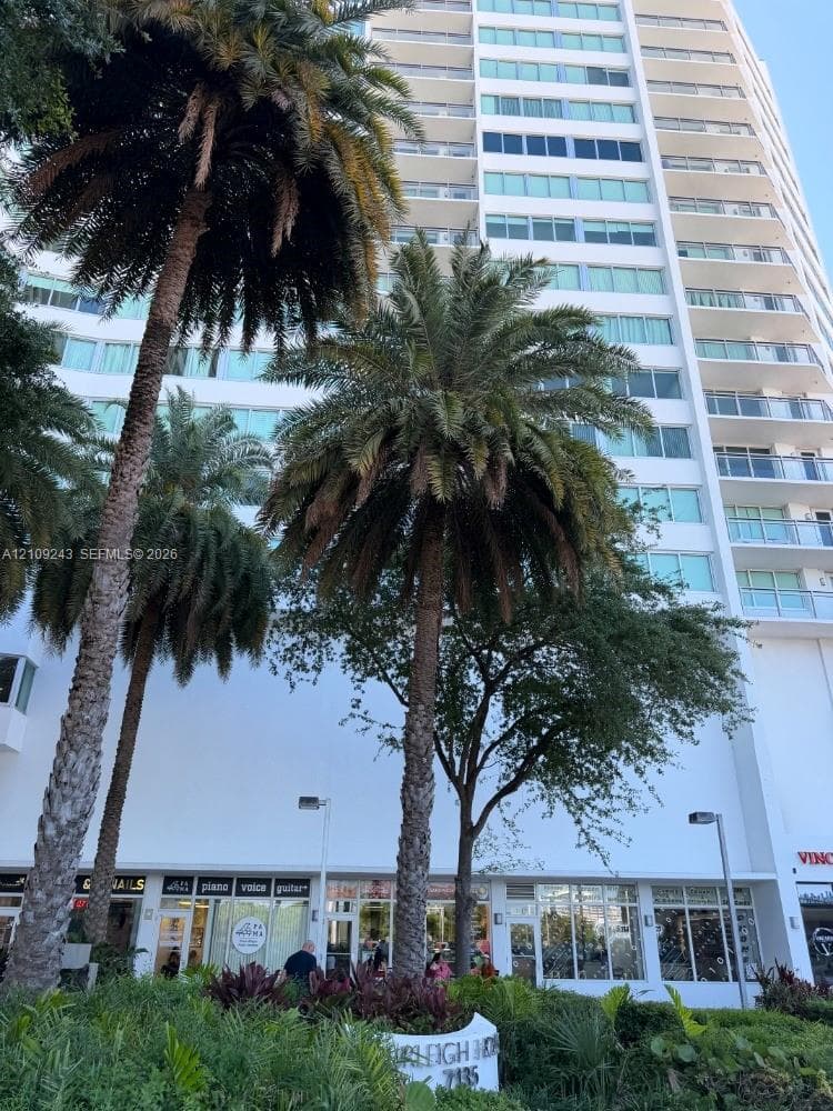 High-rise residential building exterior featuring a white façade, multiple levels of private balconies with glass railings, and ground-level commercial storefronts nestled among mature palm trees and manicured landscape planters