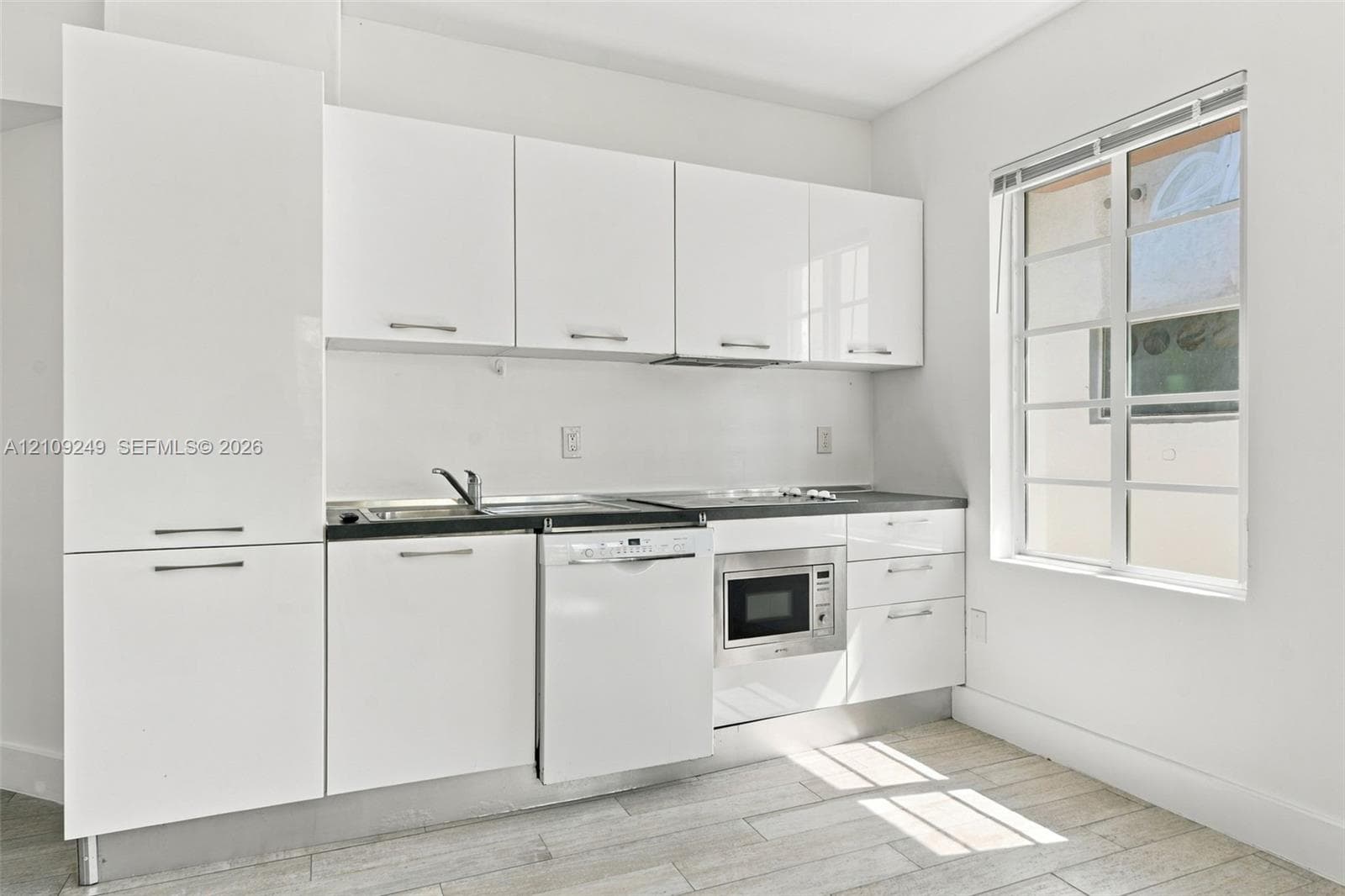 Kitchen featuring high-gloss white flat-panel cabinetry, dark countertop, integrated dishwasher, built-in microwave, and wood-finish flooring