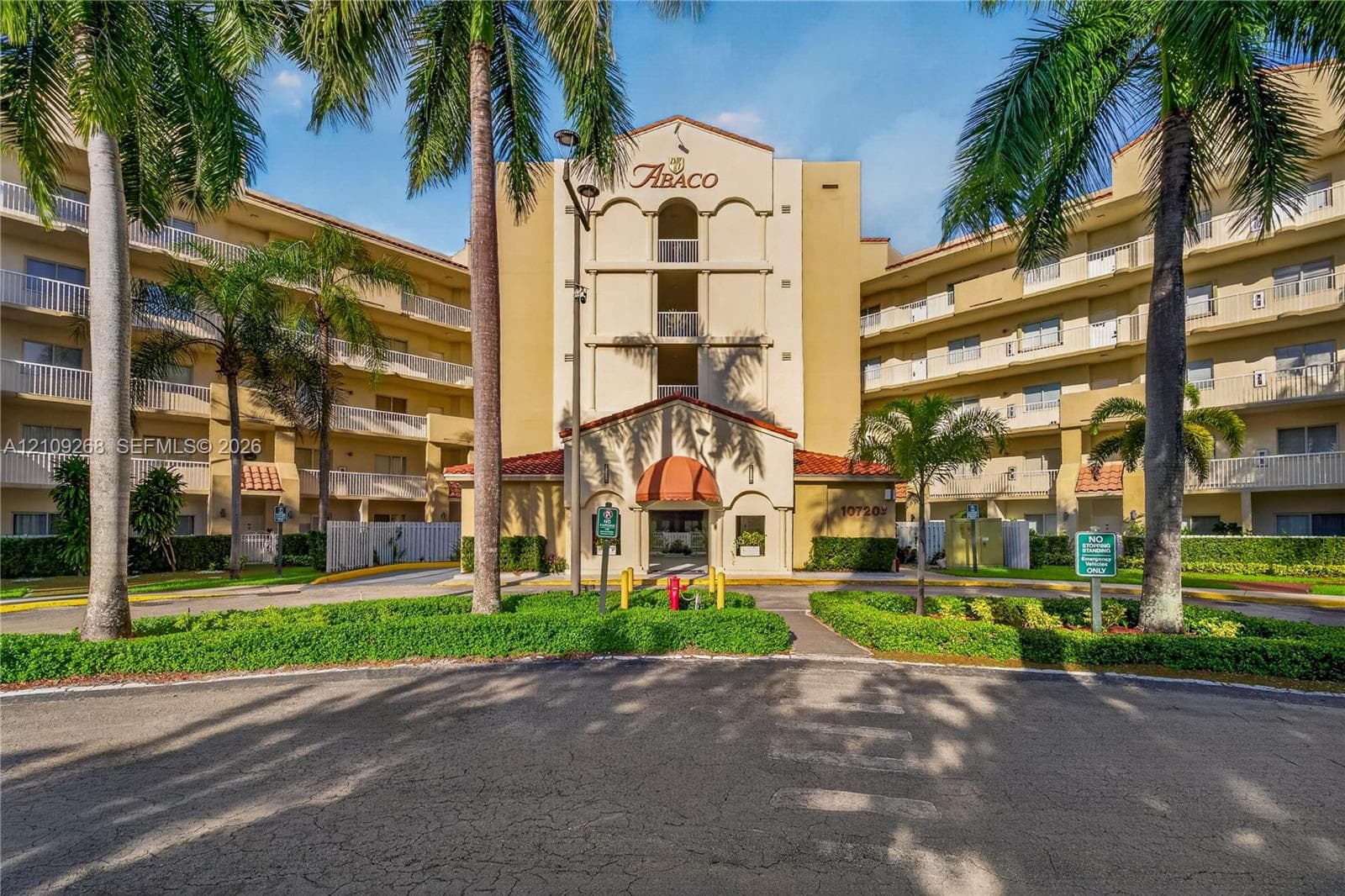 Multi-story condominium exterior featuring stucco facade, arched architectural detailing, tile roof accents, covered entryway with awning, and manicured tropical landscaping