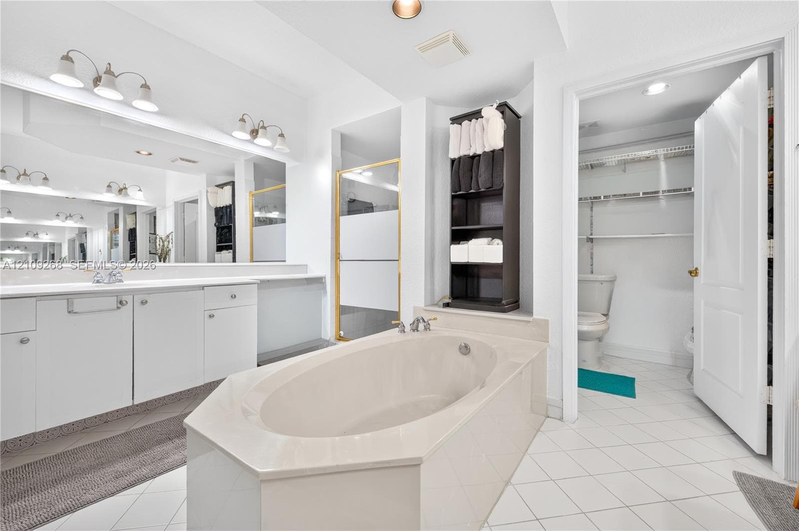 Large soaking tub with a solid surface surround, expansive wall-to-wall mirror, dual-light vanity fixtures, and white tile flooring
