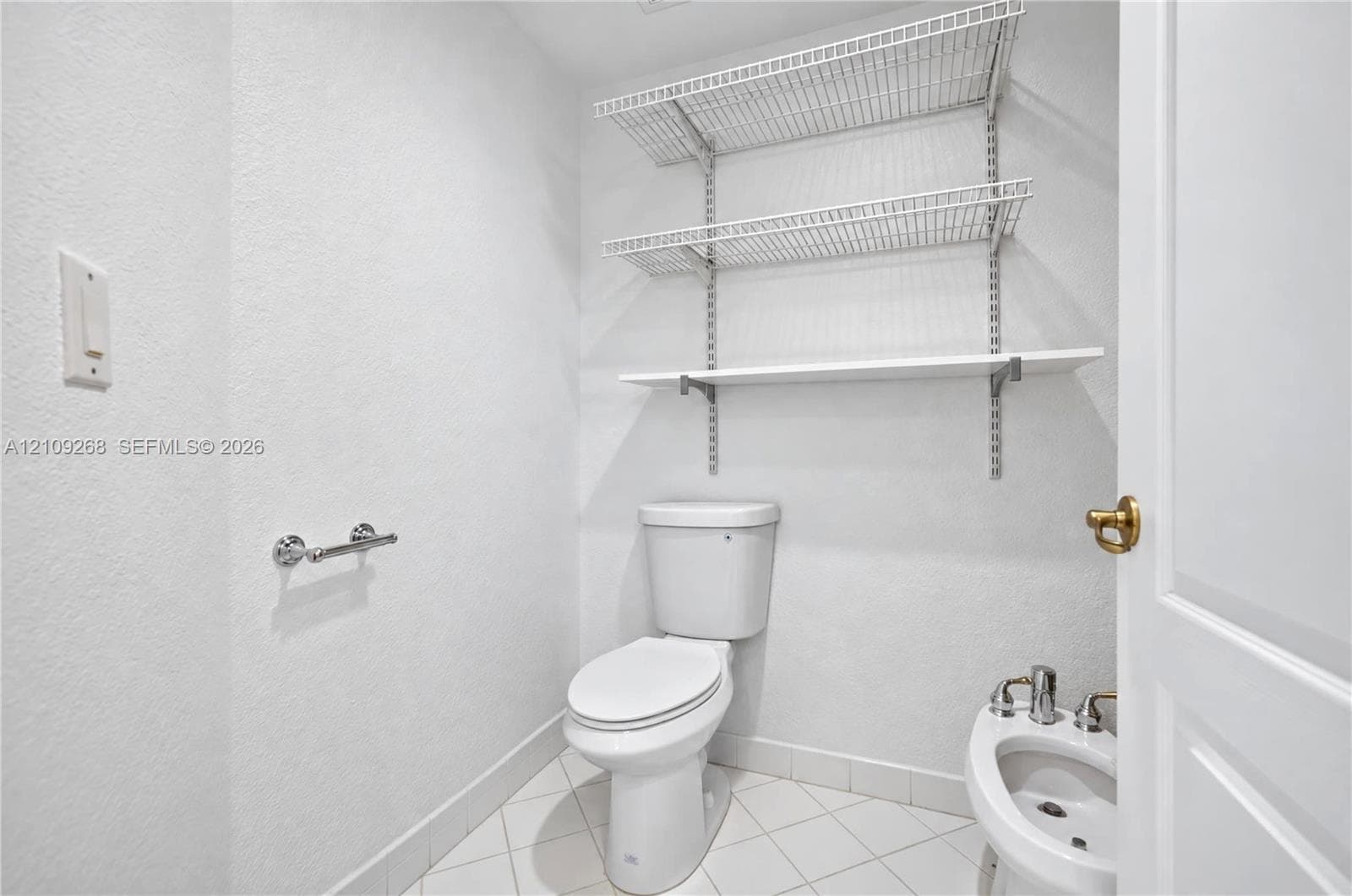 White porcelain toilet, bidet with metal hardware, tiled flooring, and adjustable wall-mounted wire shelving