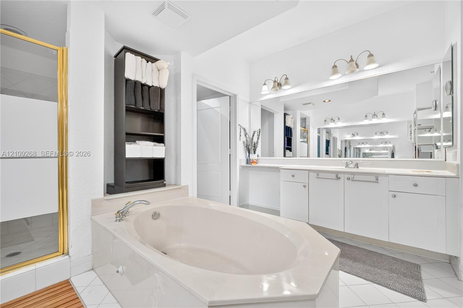 Oval soaking tub with tiled surround, glass-enclosed shower with gold-finish frame, dual-sink vanity with white cabinetry, wall-to-wall mirror, and overhead multi-bulb lighting fixtures