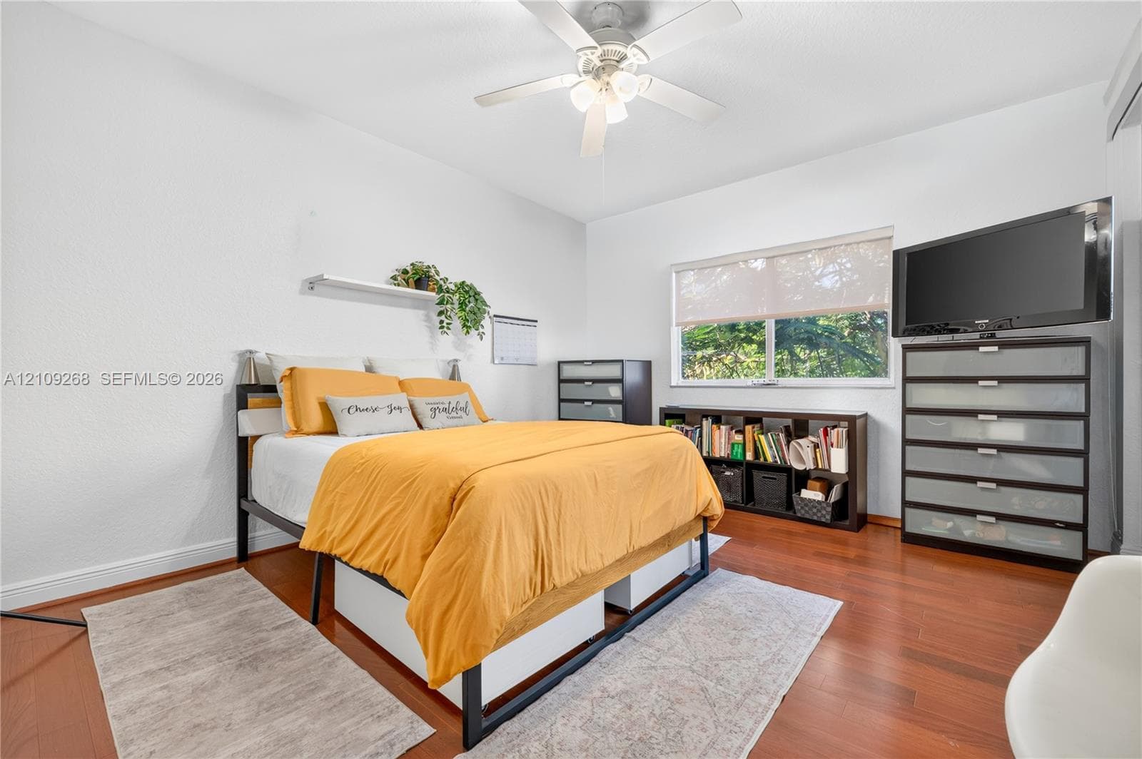 Room featuring wood-finish flooring, a ceiling fan with integrated lighting, and a window with a roller shade
