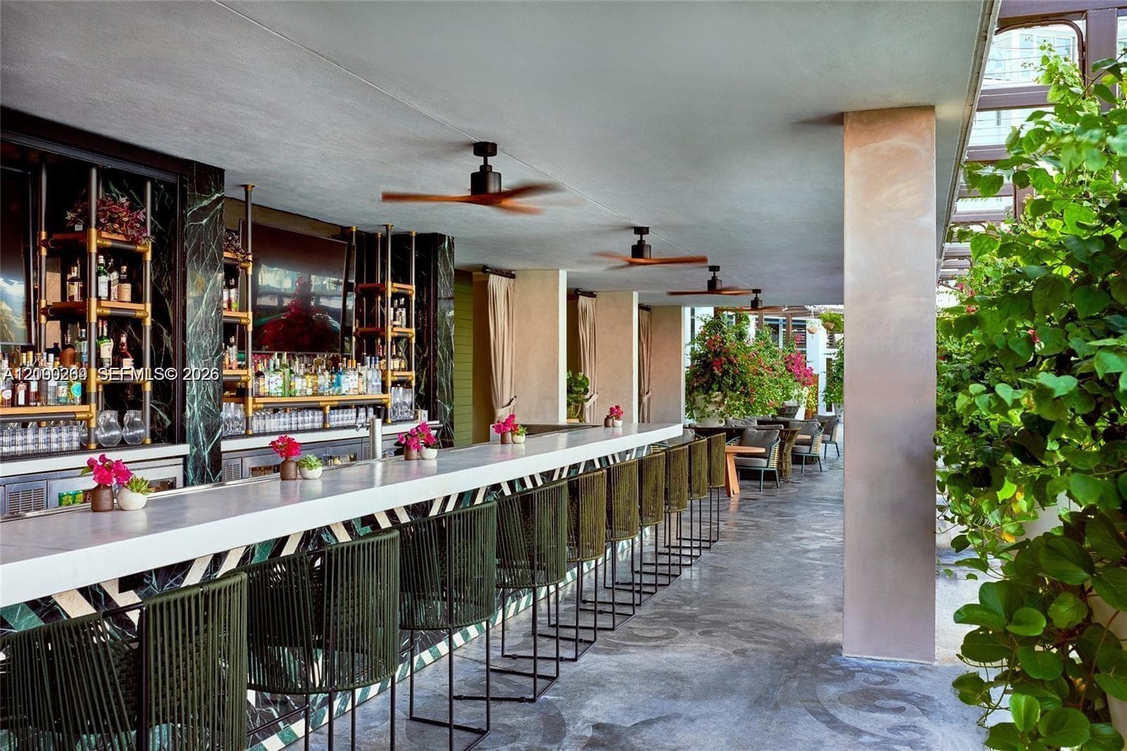 Outdoor bar area featuring an elongated bar with a solid surface countertop, built-in shelving with integrated lighting, polished stone wall cladding, and multiple overhead ceiling fans