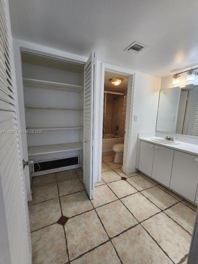 Ceramic tile flooring with decorative inlay accents, vanity with integrated basin, large wall-mounted mirror, three-light fixture, and built-in linen storage with louvered doors