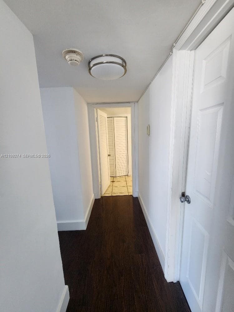 Hallway featuring dark wood-finish flooring, white paneled interior doors, a metallic flush-mount ceiling fixture, and a louvered closet door