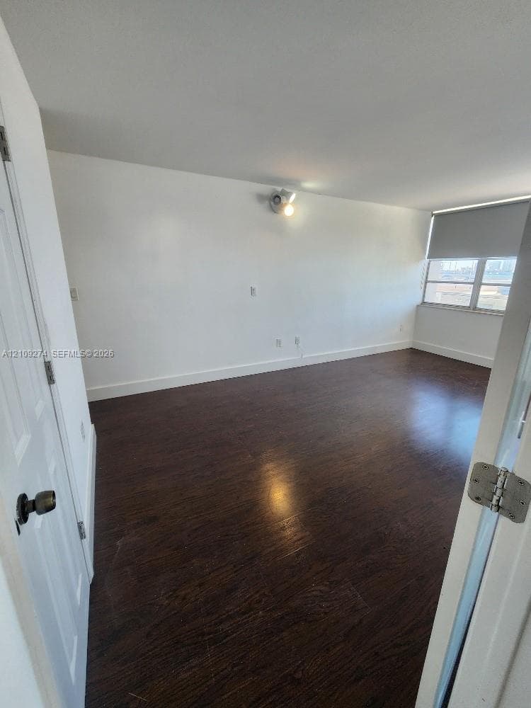 Interior room featuring dark wood-finish flooring, a wall-mounted directional light fixture, and a large window with a roller shade
