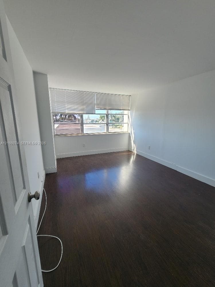 Room featuring dark-toned wood-finish flooring, white walls, and a multi-pane window with adjustable blinds