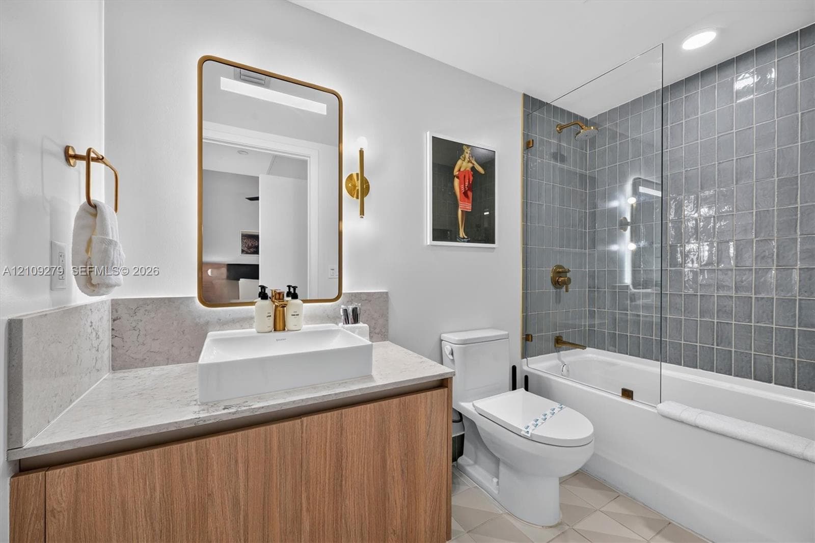 Floating wood-finish vanity with a rectangular vessel sink and stone-finish countertop, brass-toned hardware, rectangular backlit mirror, and a bathtub with gray tiled surround and glass shower panel