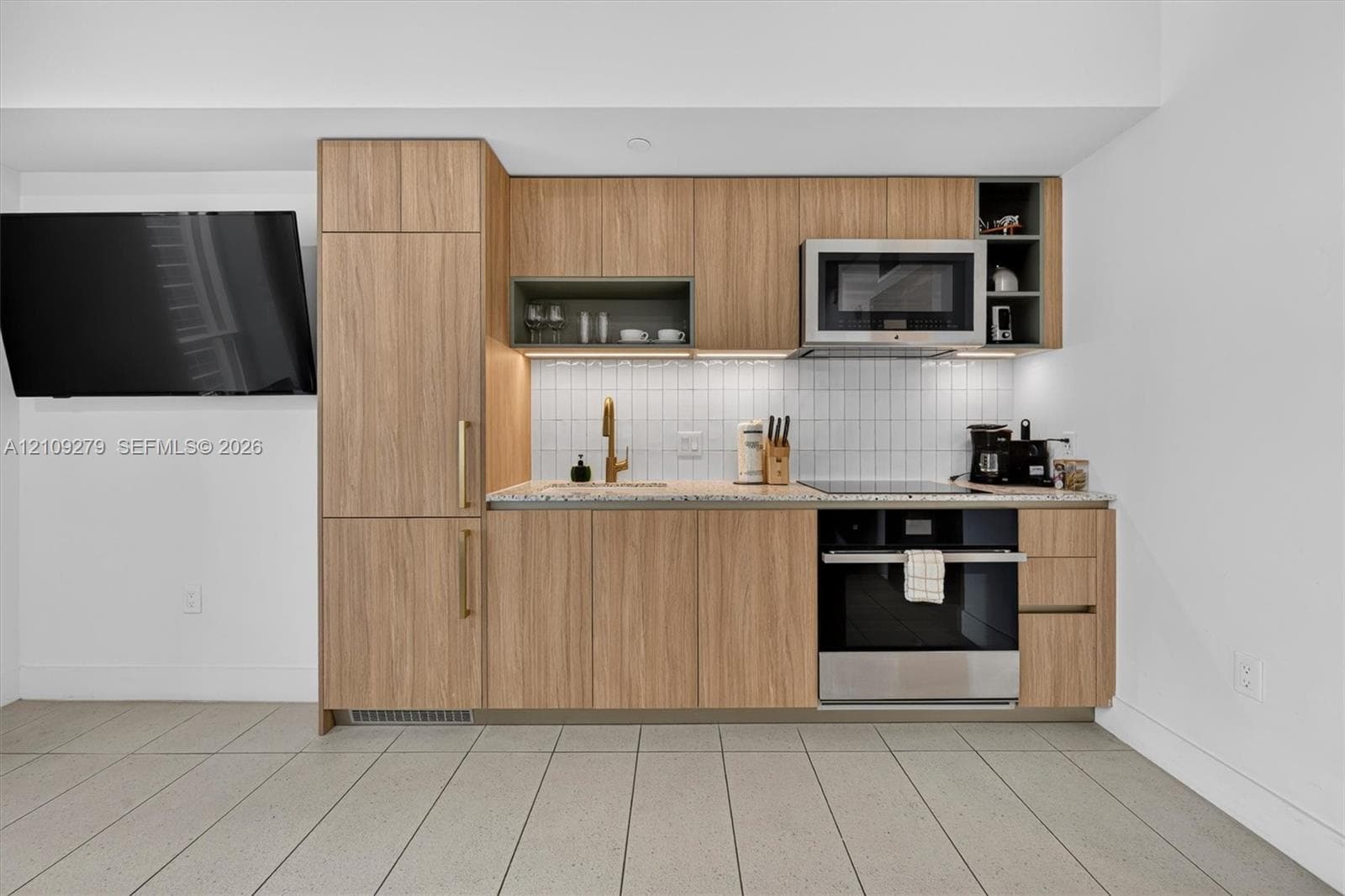 Kitchenette featuring light wood-finish cabinetry, stone-texture countertop, white subway tile backsplash, stainless steel oven and microwave, and brass-tone hardware