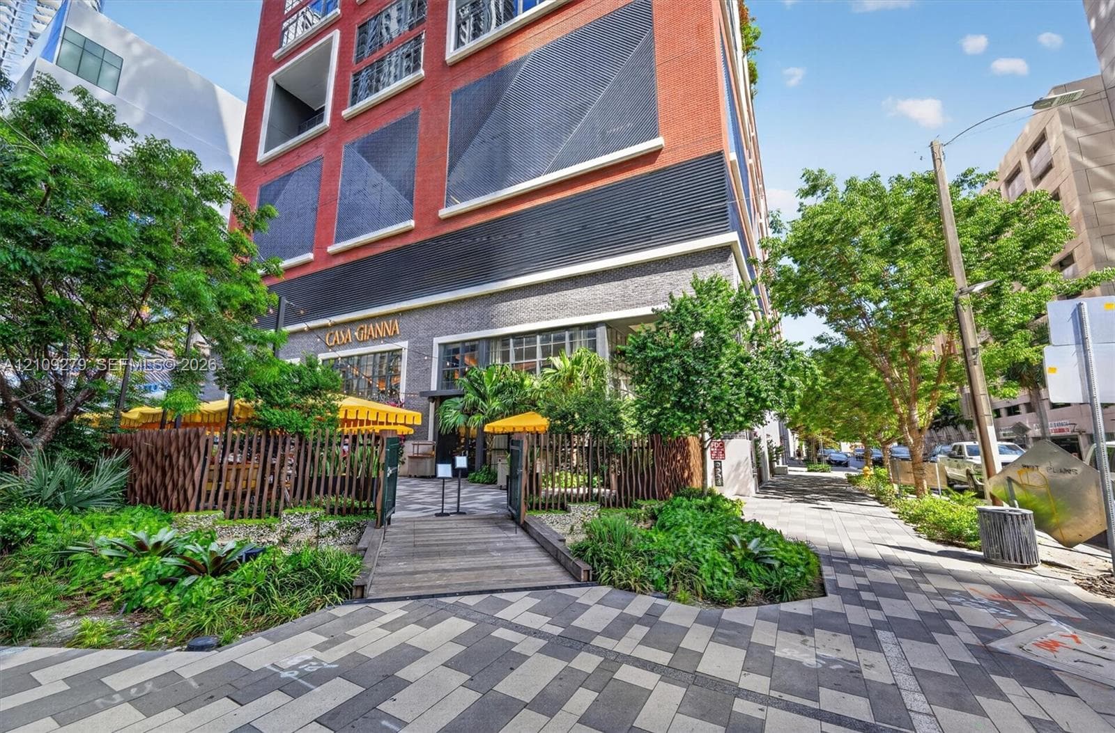 Multi-story brick exterior featuring metallic window panels, ground-level stone masonry, a elevated wood plank walkway, and integrated perimeter wood fencing