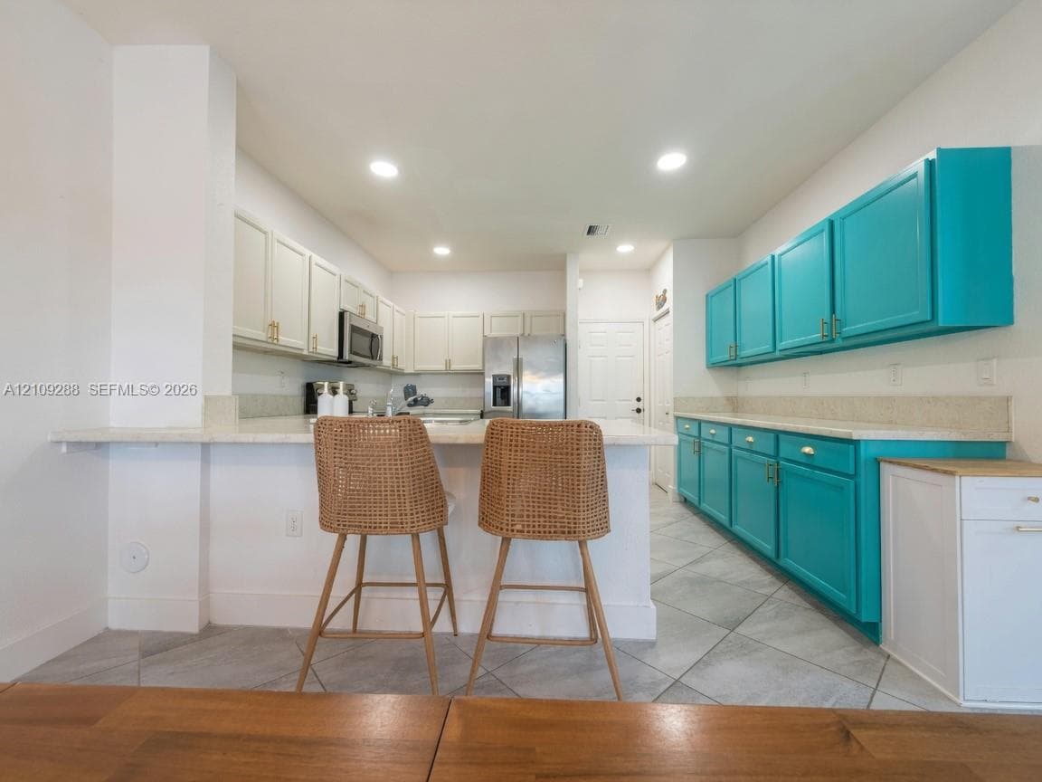 Kitchen featuring contrasting teal and white shaker-style cabinetry, stone countertops, stainless steel appliances, and large-format tile flooring