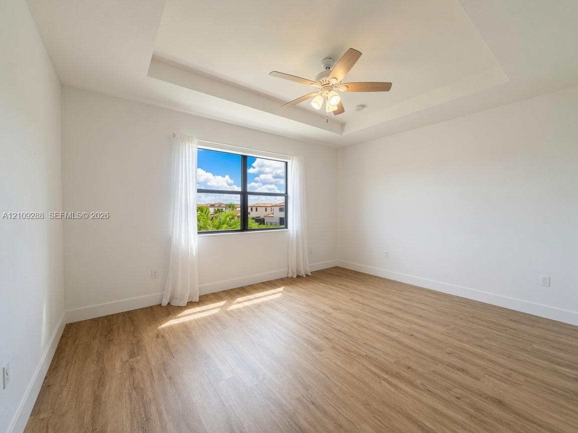 Tray ceiling with integrated lighting, wood-finish flooring, and a multi-blade ceiling fan featuring light fixtures