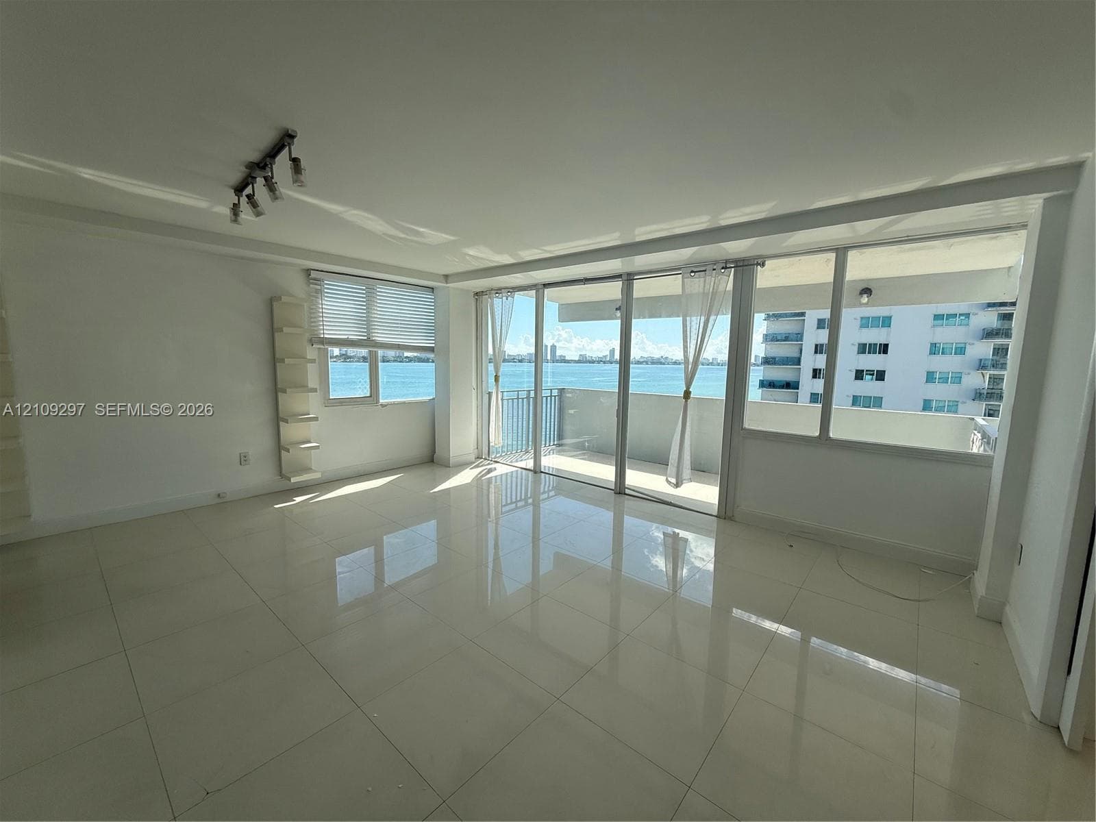 Spacious room featuring light-colored polished tile flooring, floor-to-ceiling glass sliding doors, a built-in wall-mounted shelving unit, and an attached balcony