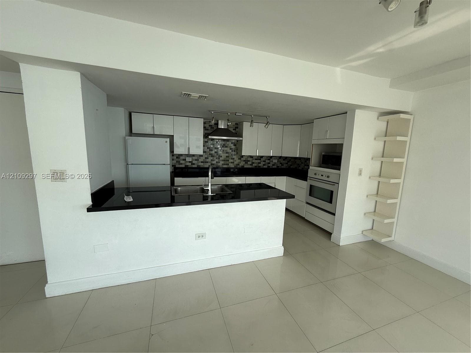 Kitchen featuring white cabinetry, dark countertops, a mosaic tile backsplash, a stainless steel dual-basin sink, and built-in shelving
