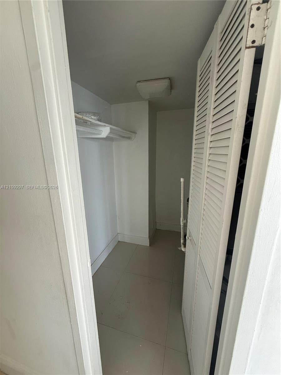 Storage closet featuring louvered bi-fold doors, white shelving, tile flooring, and a flush-mount light fixture