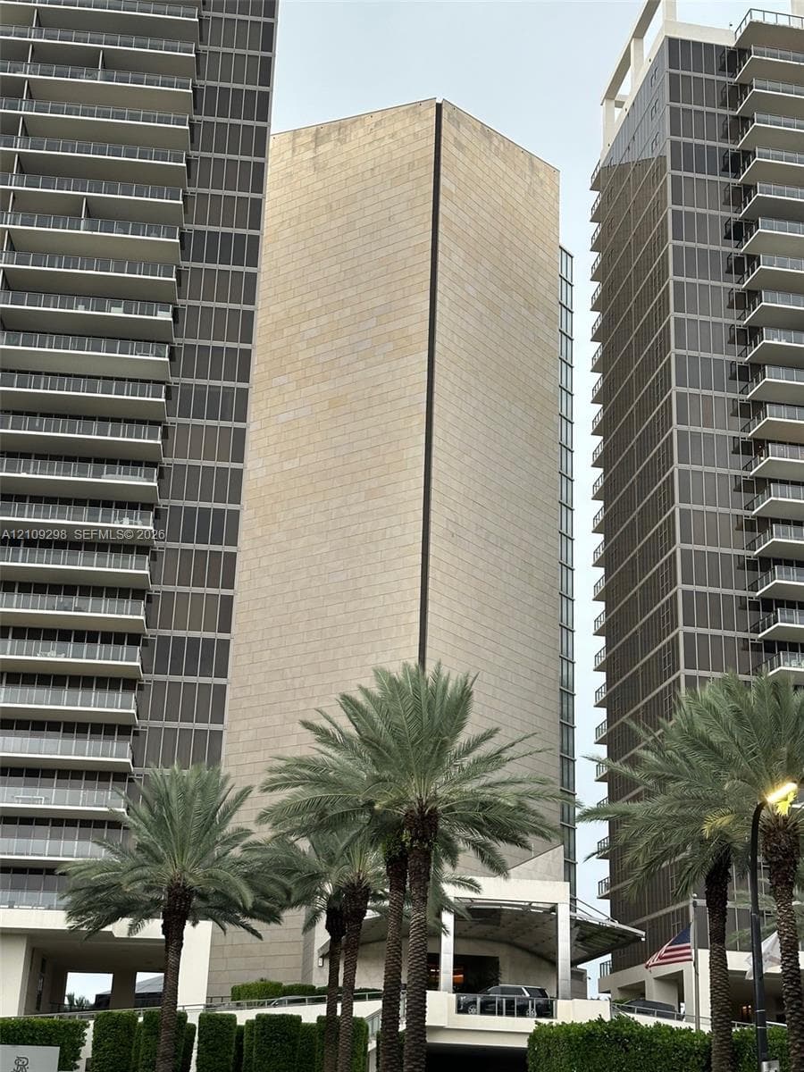 High-rise architectural exterior featuring glass-railed private balconies, paved motor court, mature palm trees, and manicured landscaping