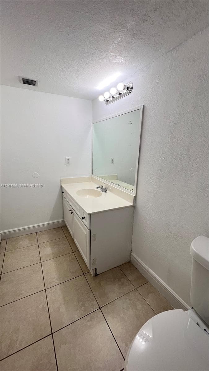 Bathroom featuring a white vanity cabinet, solid-surface integrated countertop with basin, wall-mounted mirror, four-light vanity fixture, and tiled flooring