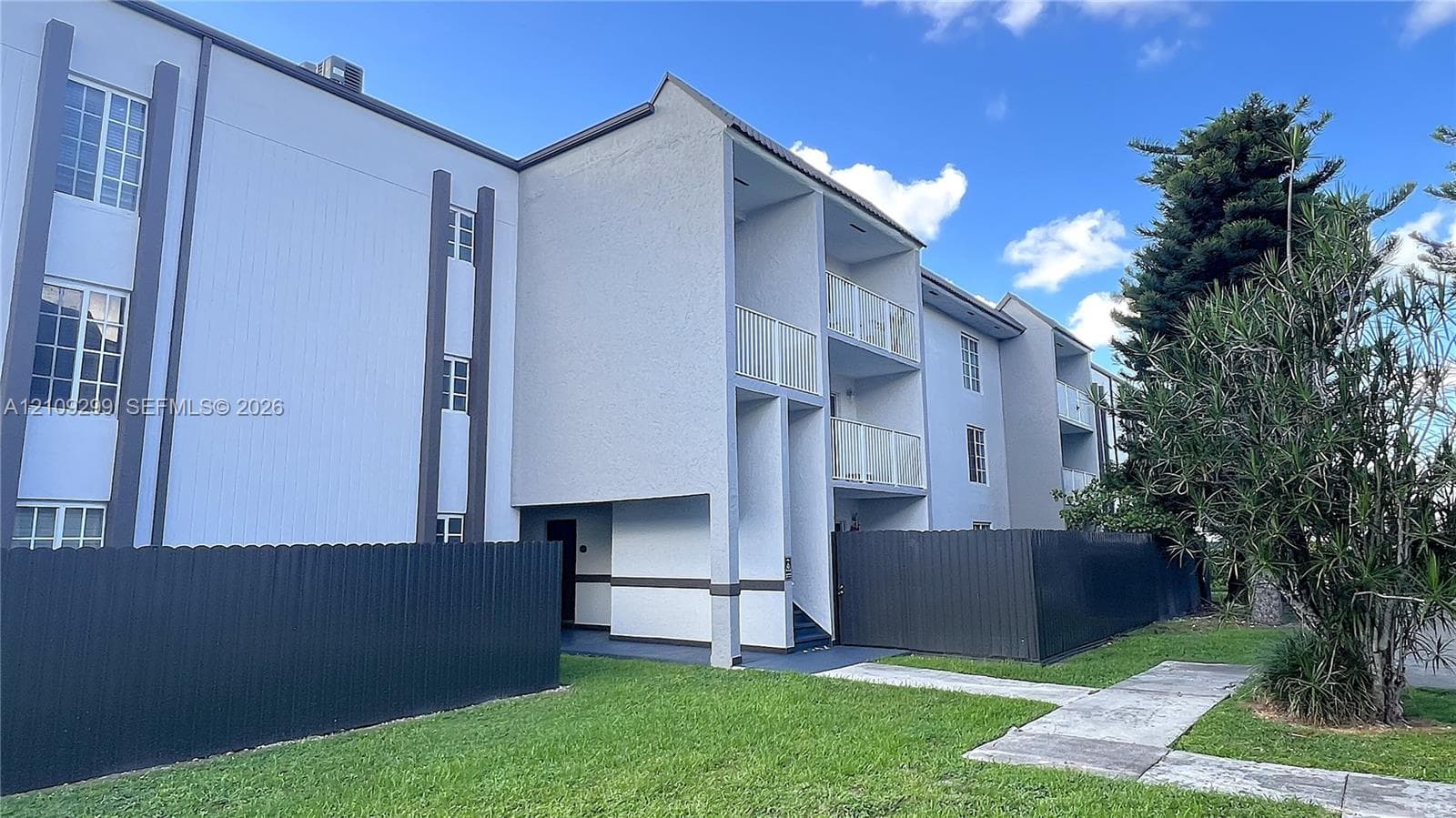 Multi-story exterior featuring a light-toned stucco façade, private elevated balconies with metal railings, and dark-finished metal privacy fencing