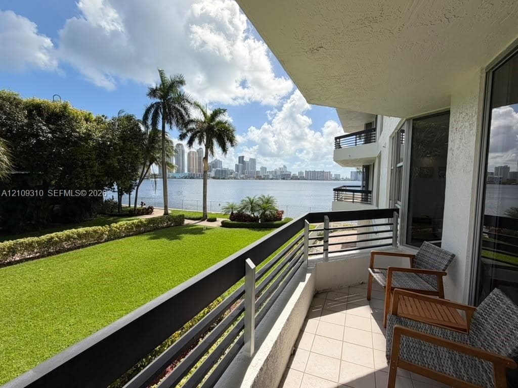 Covered balcony featuring tile flooring and horizontal metal railings, overlooking a waterfront lawn, palm trees, and the city skyline